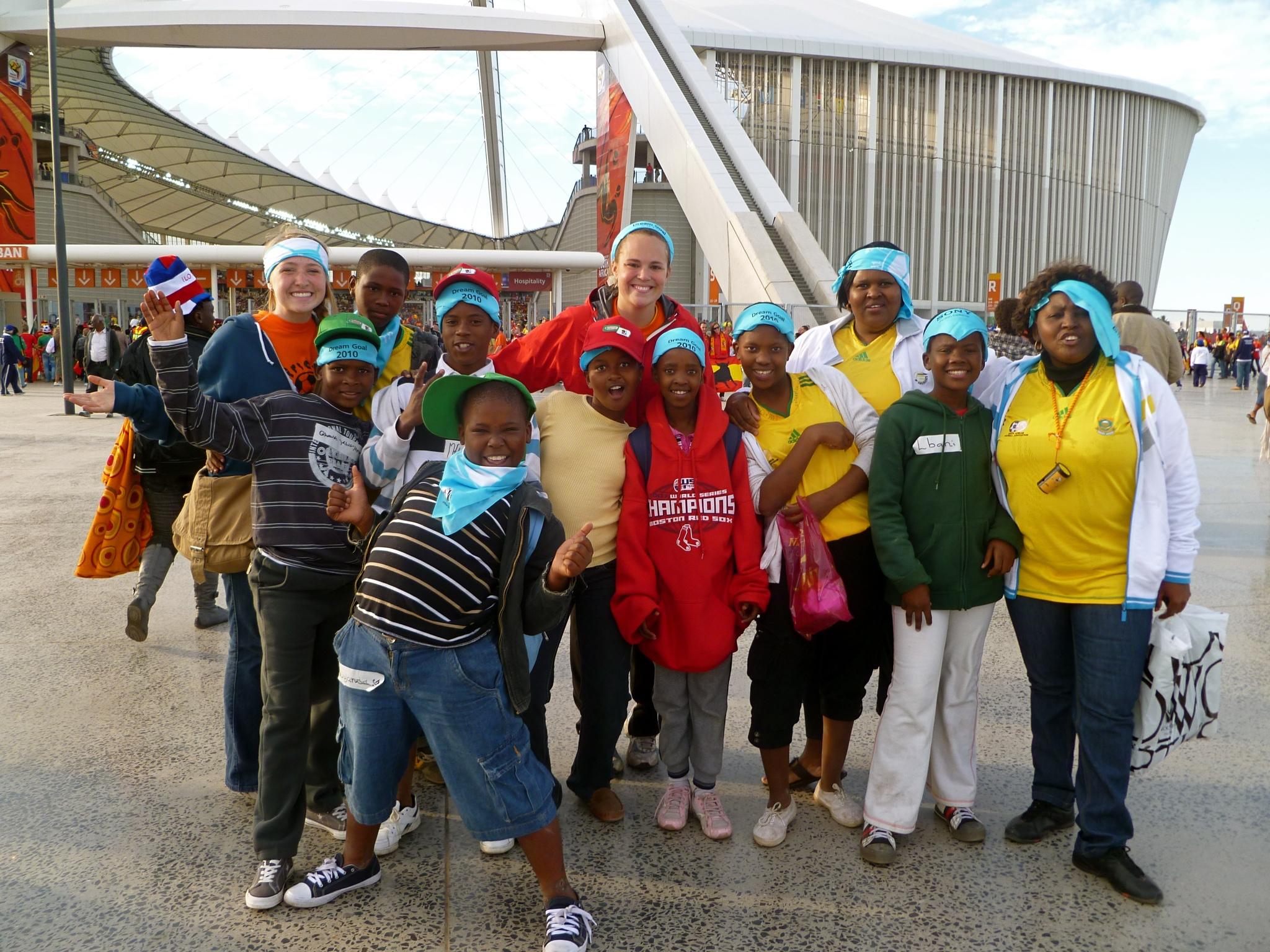 A blonde American girl stands with several Black children in front of a large soccer stadium.