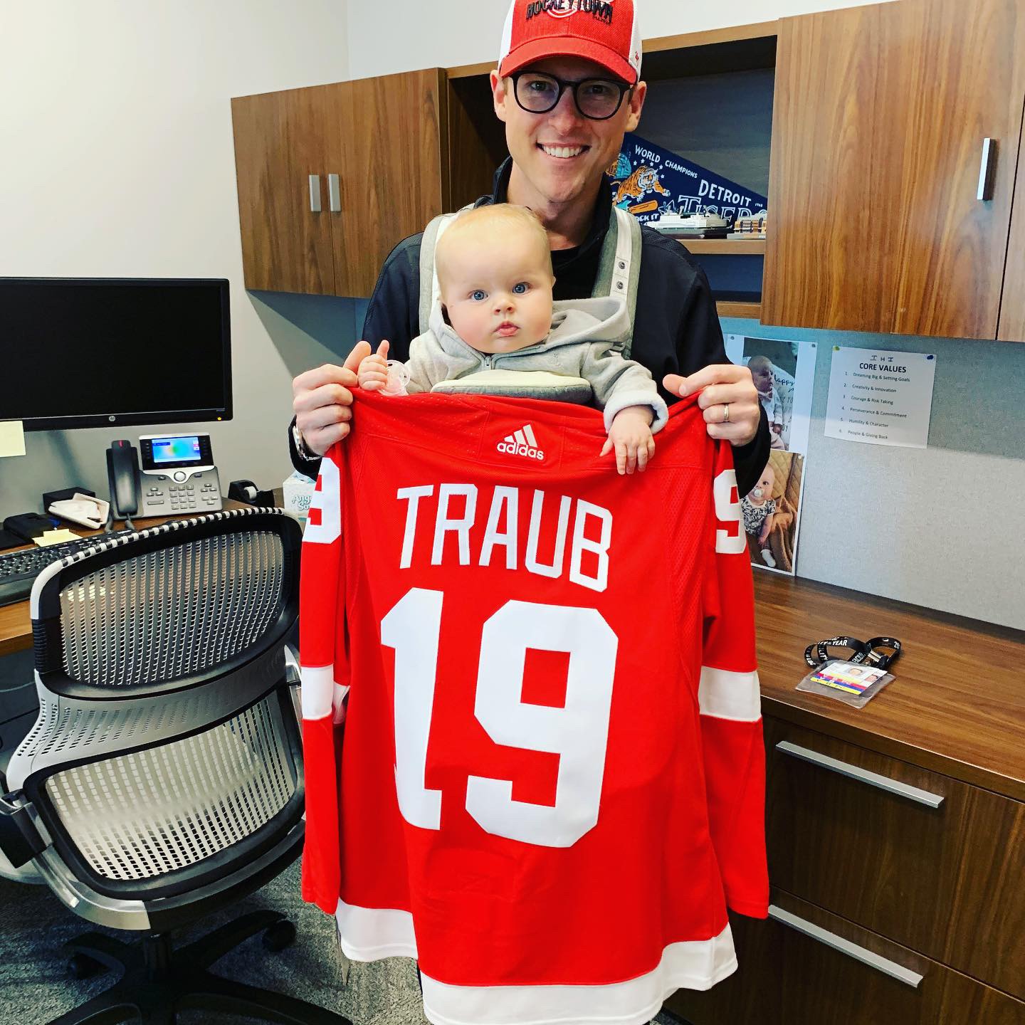 A person with a baby in a front-facing baby carrier holds up a red and white hockey jersey with the name "Traub" on the back.