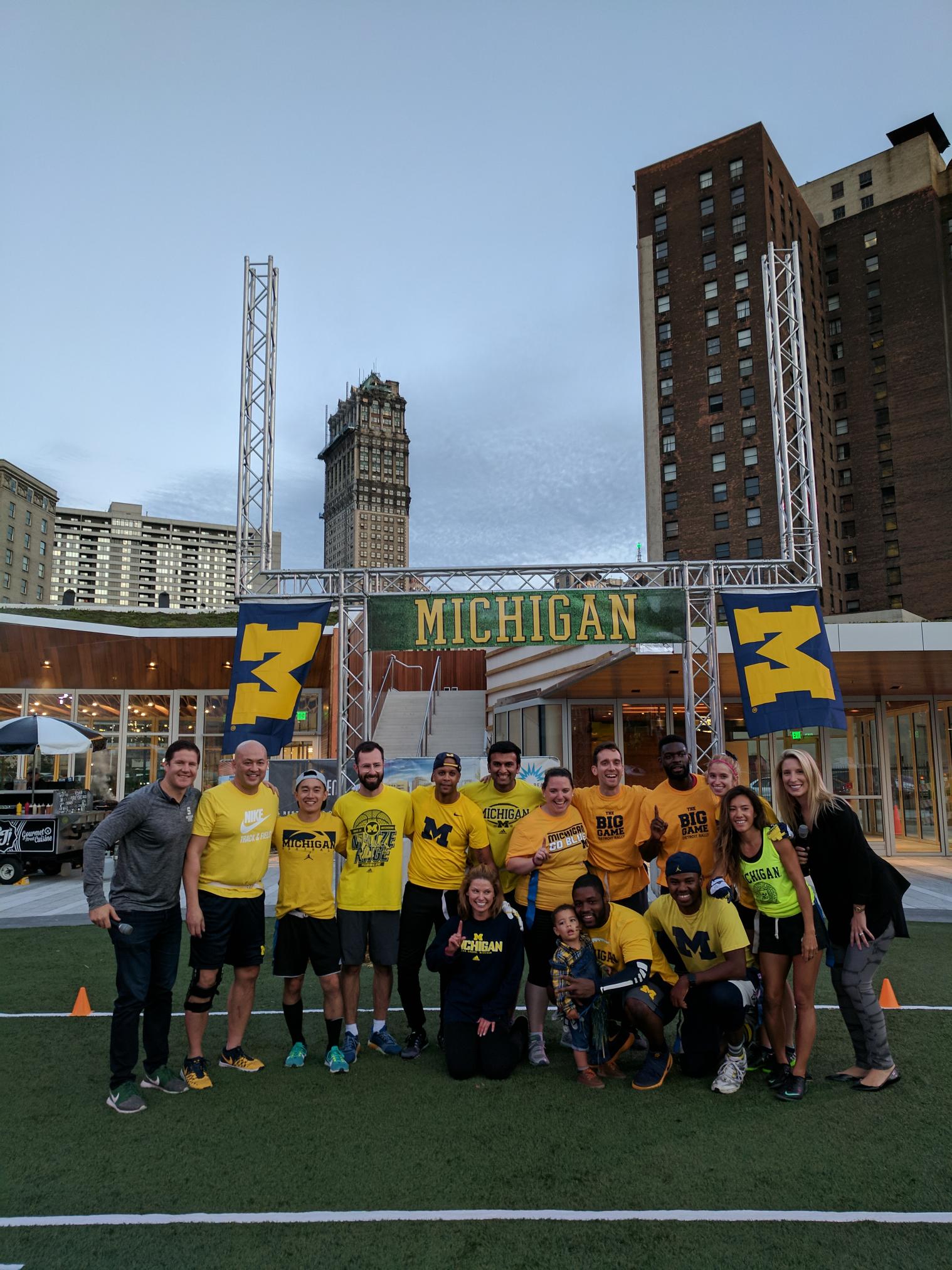 A small group of people in University of Michigan shirts gather in front of a pair of football goal posts with U of M flags hanging off.