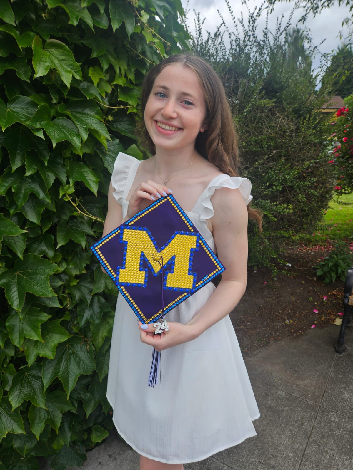 A teenage girl in a white dress holds up a graduation cap bedazzled with a blue and yellow block M.