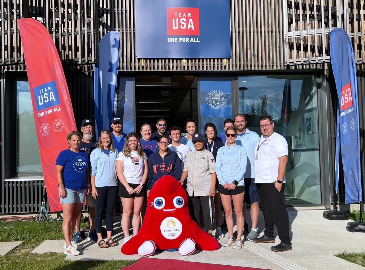 A group of people outside a building with a sign that reads "Team USA: One For All"