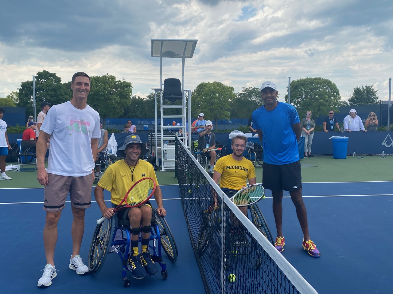 Two people in yellow Michigan shirts sit in wheelchairs holding tennis rackets next to Joe Salisbury and Rajeev Ram, a highly successful doubles team in professional tennis.