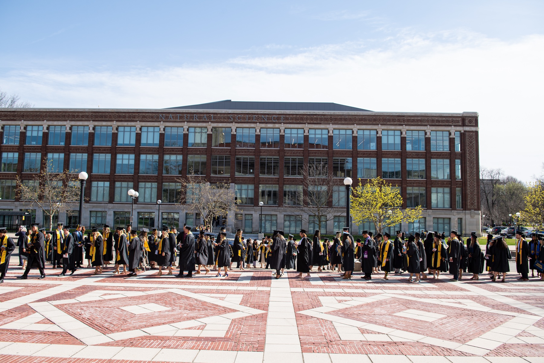 A crowd of faculty and students in commencement robes mill about in front of the School of Kinesiology Building.