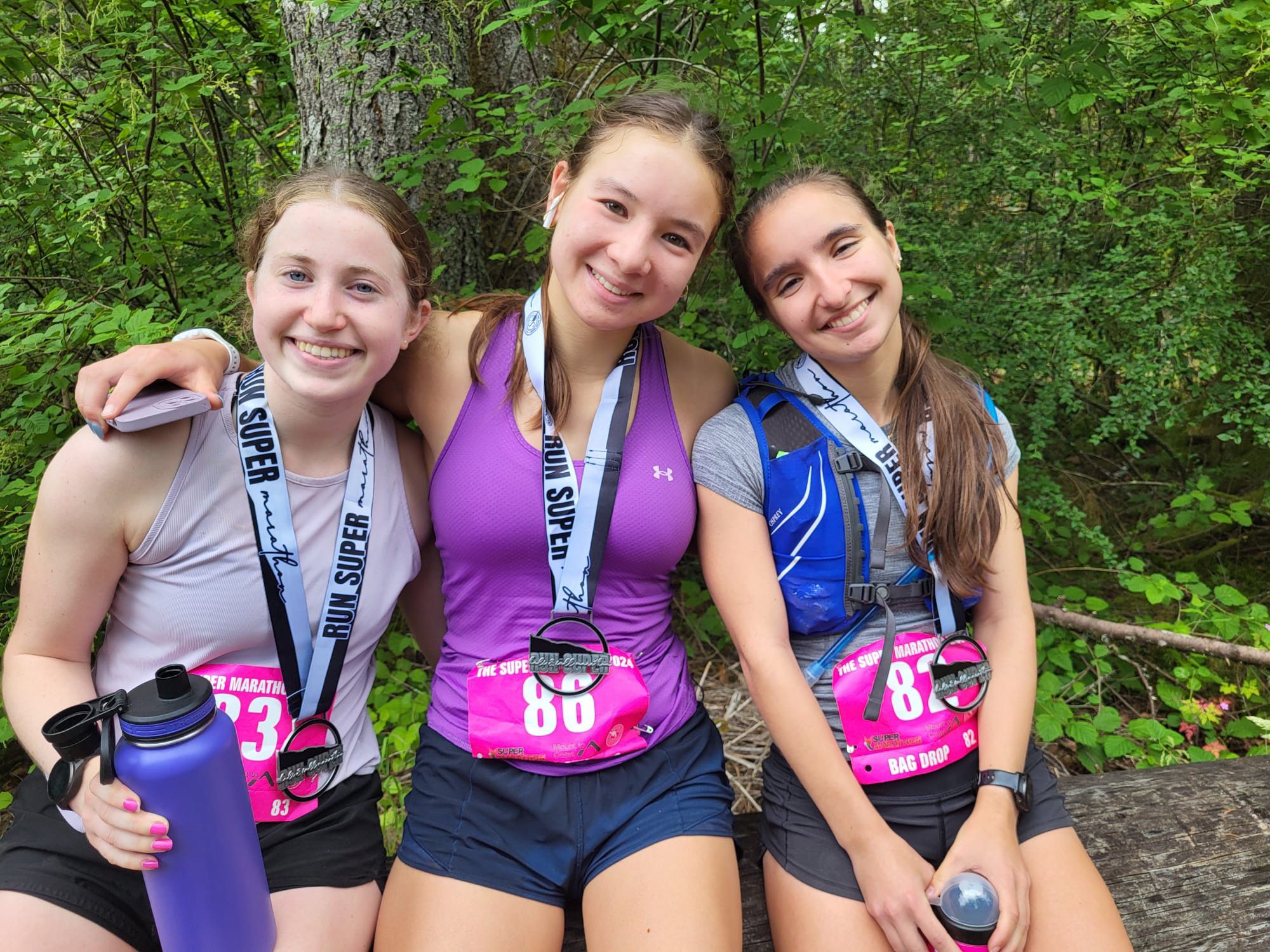 Three teenage girls sit on a log in running shorts and athletic tanks, wearing race numbers and finisher medals. Two hold water bottles.