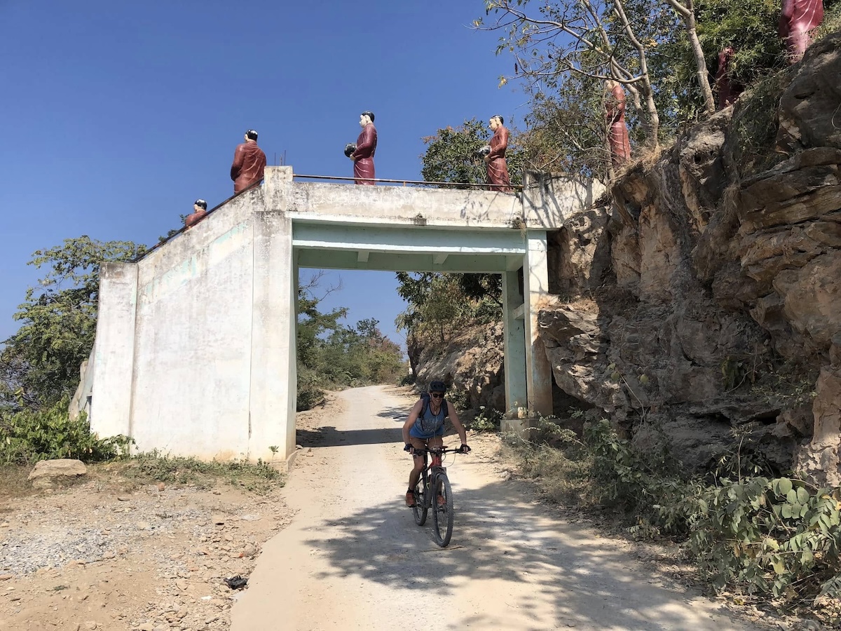 Saari mountain biking under an overpass decked with statues in Myanmar