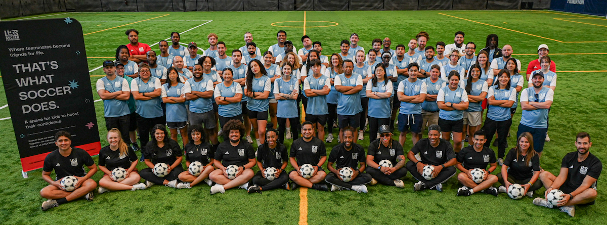 About 60 people stand with their arms crossed, wearing light blue soccer jerseys with white sleeves. In front of them, 13 people sit cross-legged with soccer balls in their laps, wearing black T-shirts.