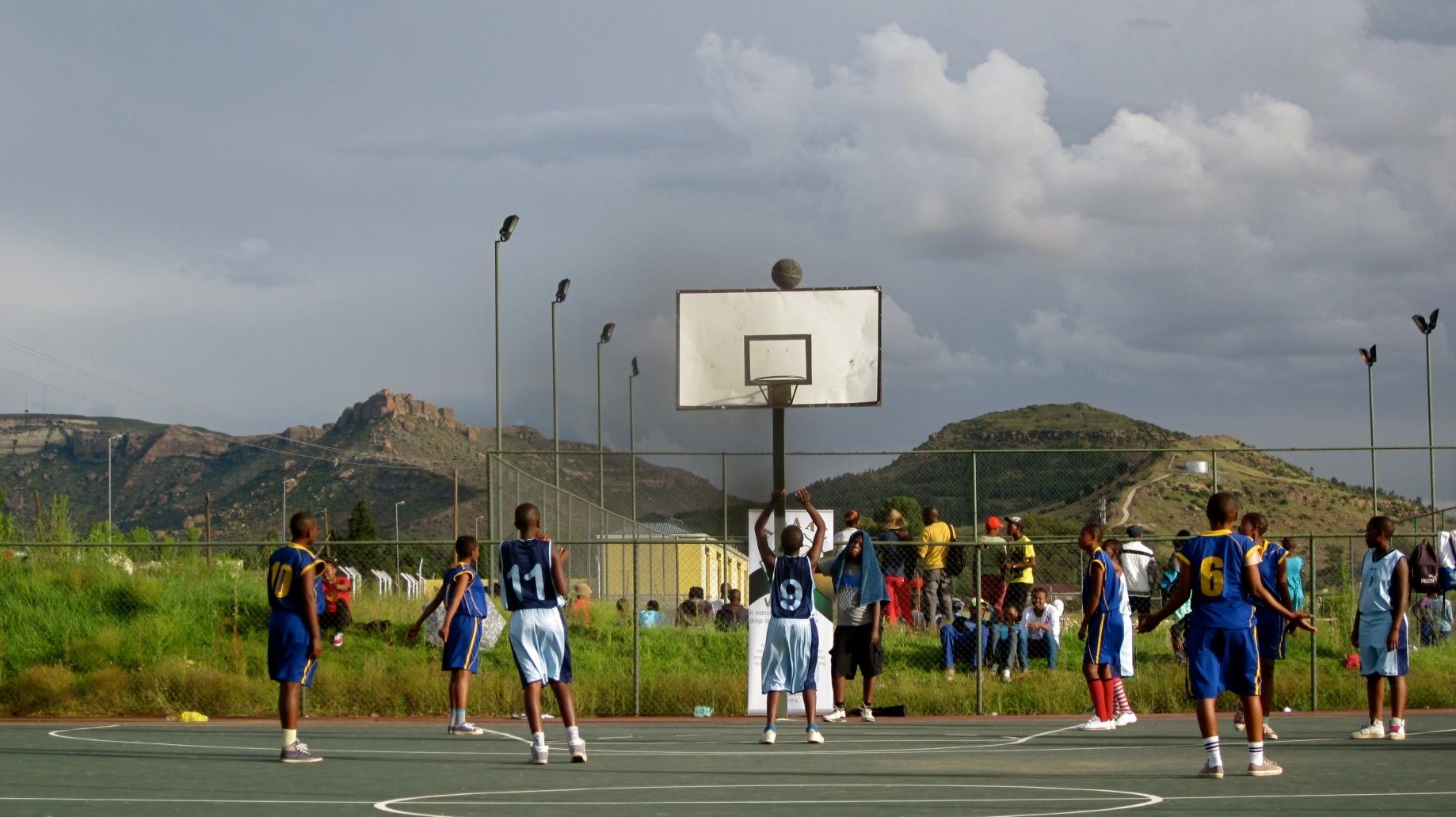 Kids in basketball uniforms play basketball on a court in front of a large mountain.