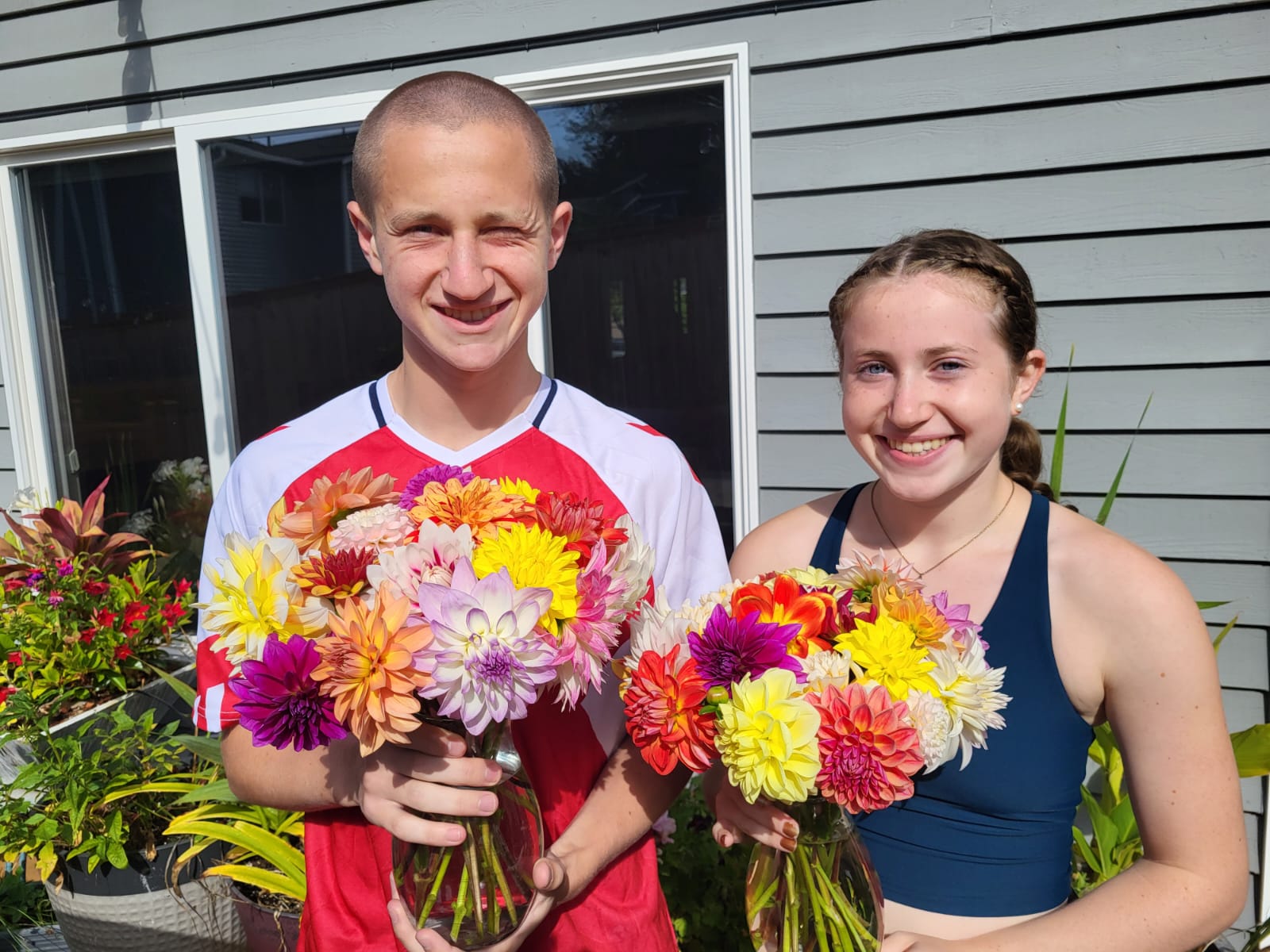 Two teenagers stand outside a grey house holding bouquets of brightly colored flowers.