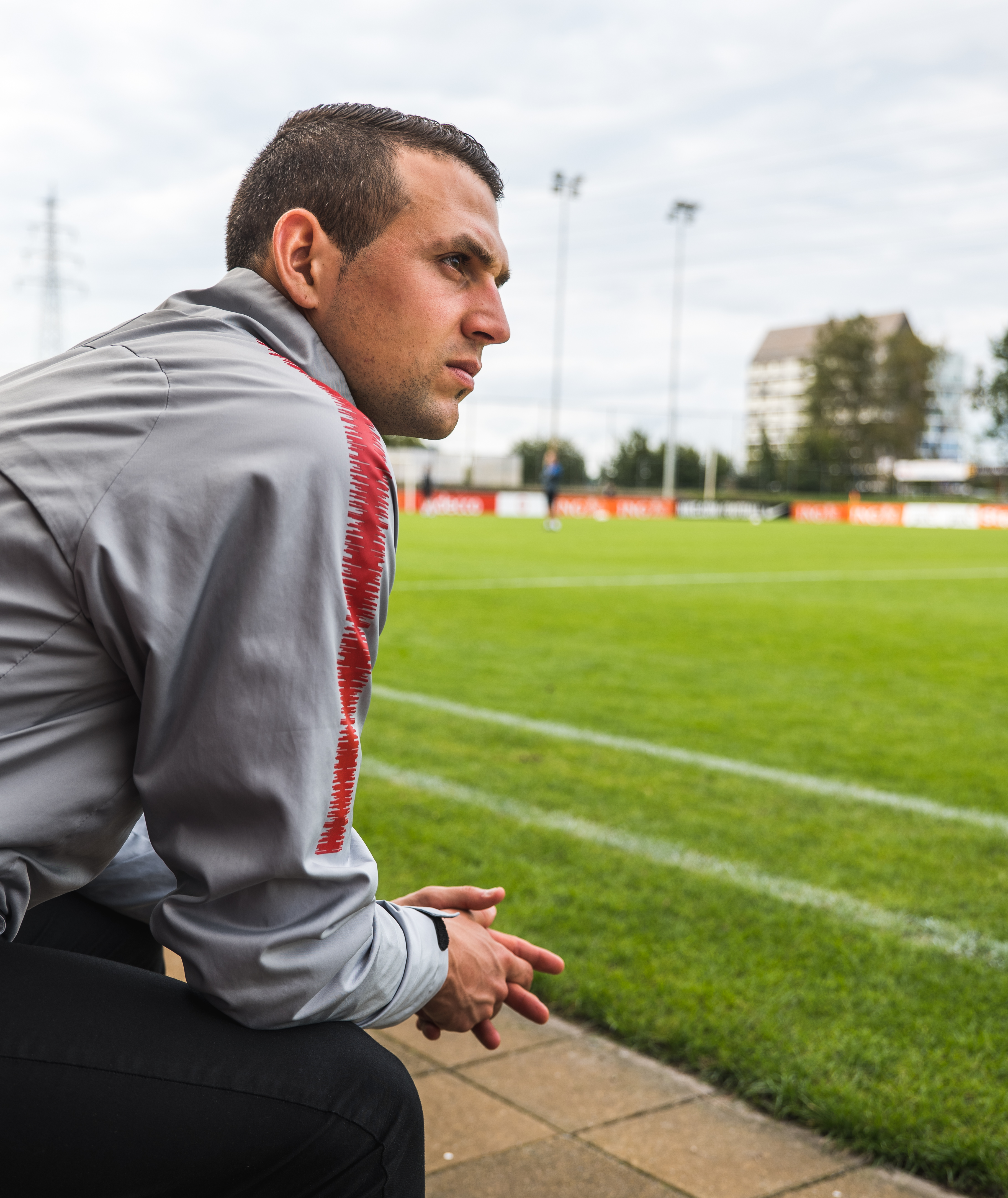 Peri Marosevic sitting on the sidelines of a soccer field in a grey warm-up jacket