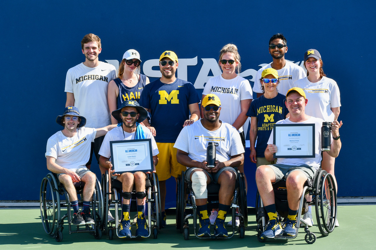 A group of people in white, blue, and yellow Michigan gear smile at the camera. Two hold plaques announcing they were named All-Americans.