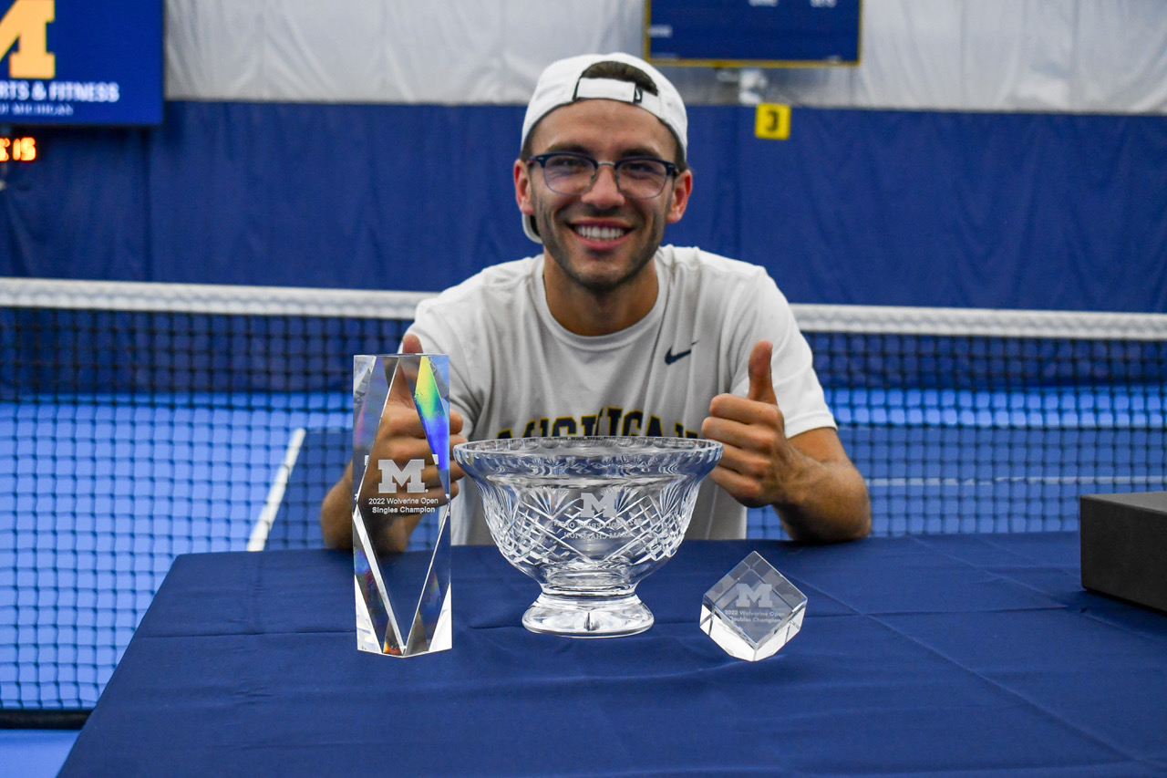 A person in a white shirt, backward baseball cap, and glasses gives two thumbs up behind a table with several trophies on it. He's sitting on a tennis court.