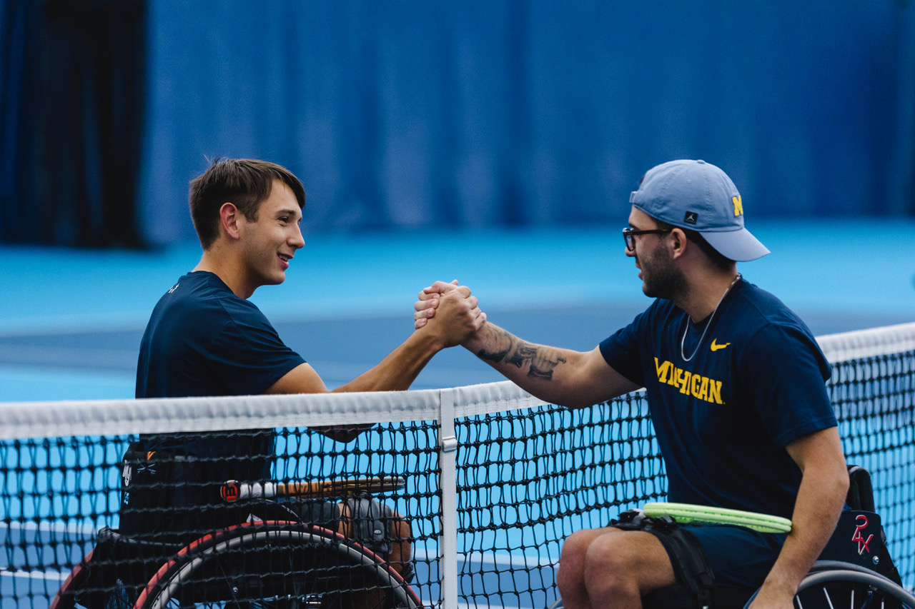 Two people in navy blue shirts and wheelchairs shake hands over a tennis net.